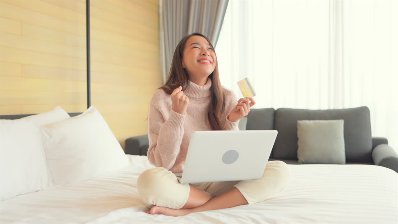 예쁜 젊은 여성이 침대에 앉아 손에 신용카드를 들고 노트북에 입력한 다음 성공적인 온라인 구매 경쟁에 반응합니다.