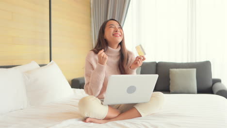 A-pretty-young-woman,-sitting-on-a-bed,-credit-card-in-hand-hits-enter-on-her-laptop,-then-reacts-to-her-successful-competition-of-an-online-purchase