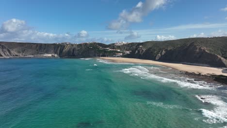 Aerial-shot-flying-over-a-beautiful-bay-with-turquoise-water-towards-houses,-surrounded-by-cliffs-and-green-hills-on-a-sunny-day