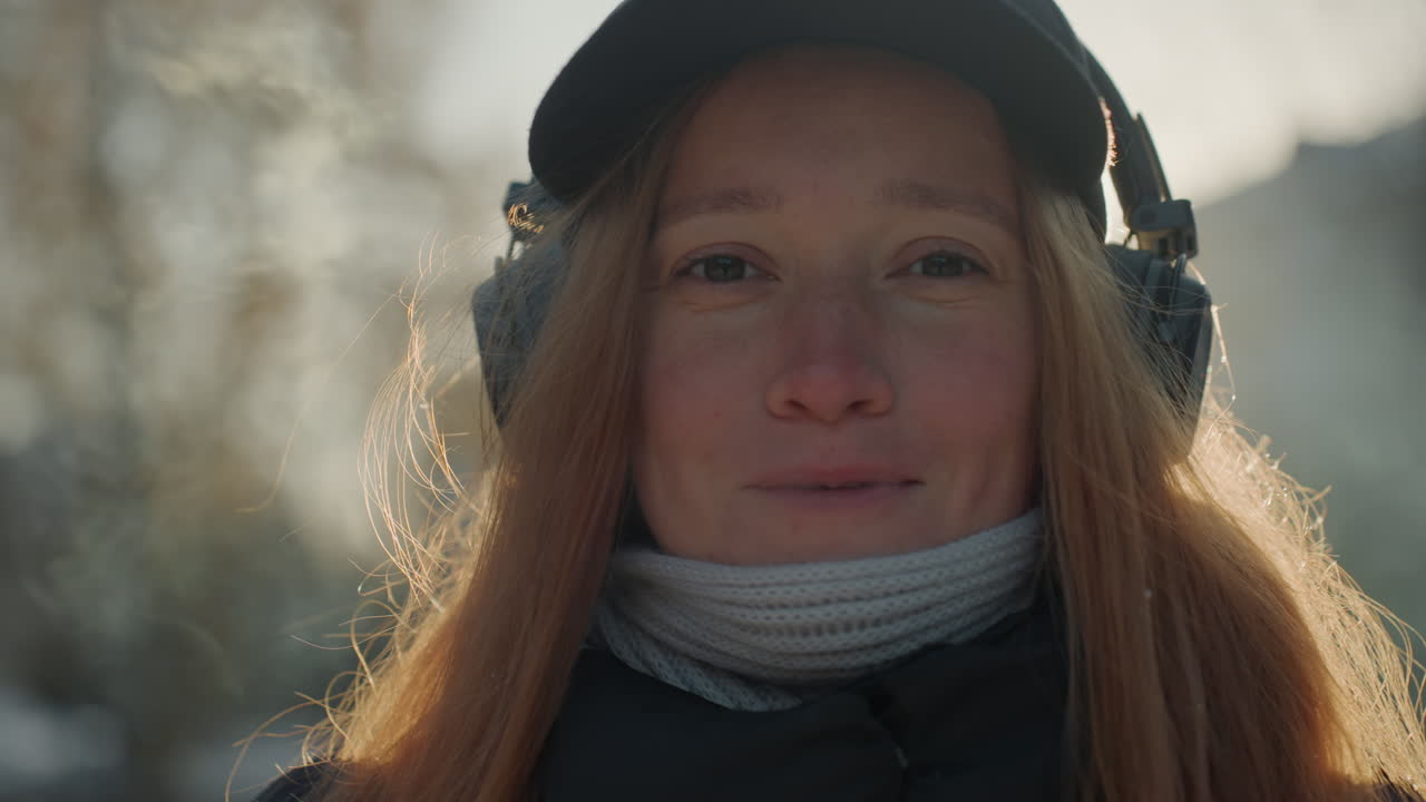 White woman with headphones smiling in sunrise backlight, warm golden glow on red hair, candid urban closeups with scarf and cap, gentle hopeful expression, soft bokeh city trees, relaxed commuter