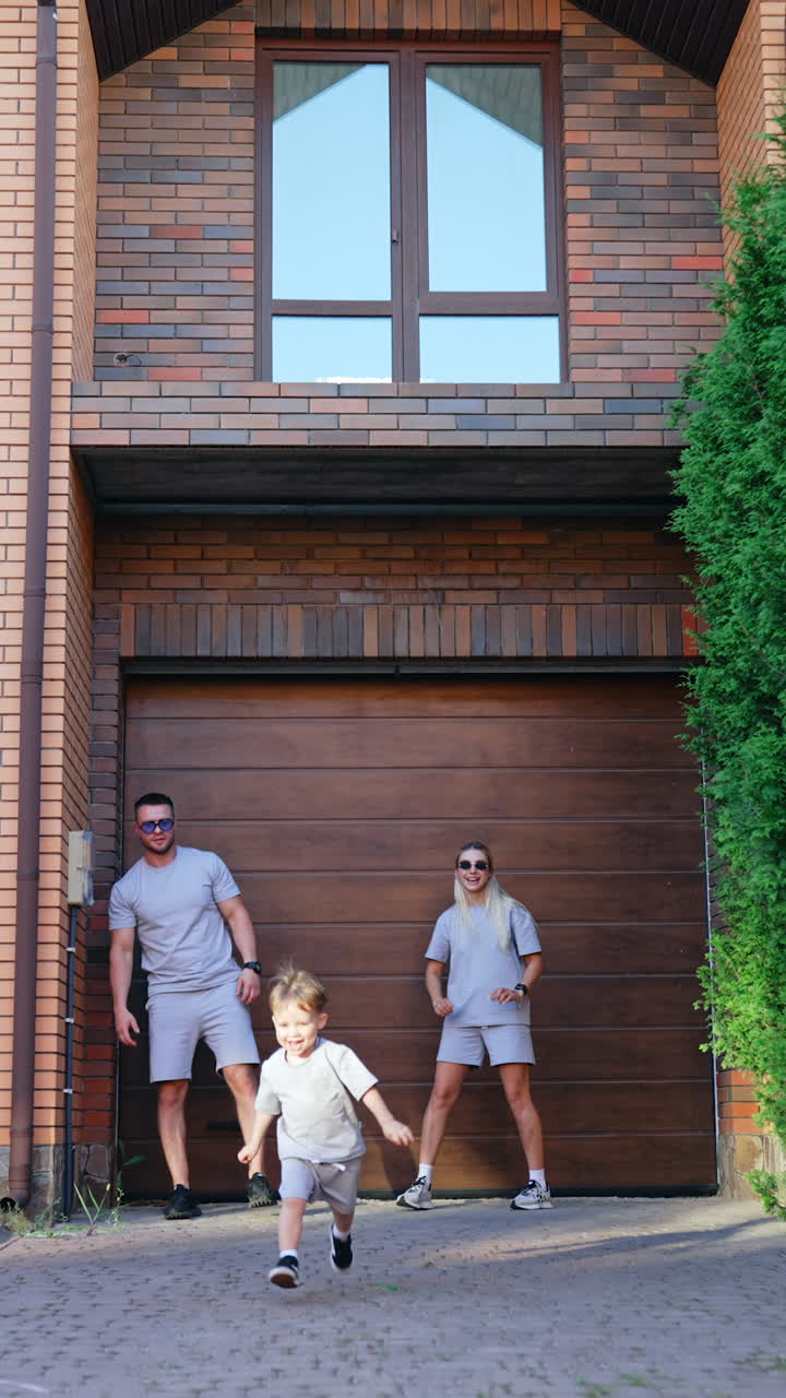 Familie in bijpassende outfits buiten. Een gezin van drie speelt buiten hun huis, met een kind dat vrolijk rent in casual bijpassende outfits bij een garage.