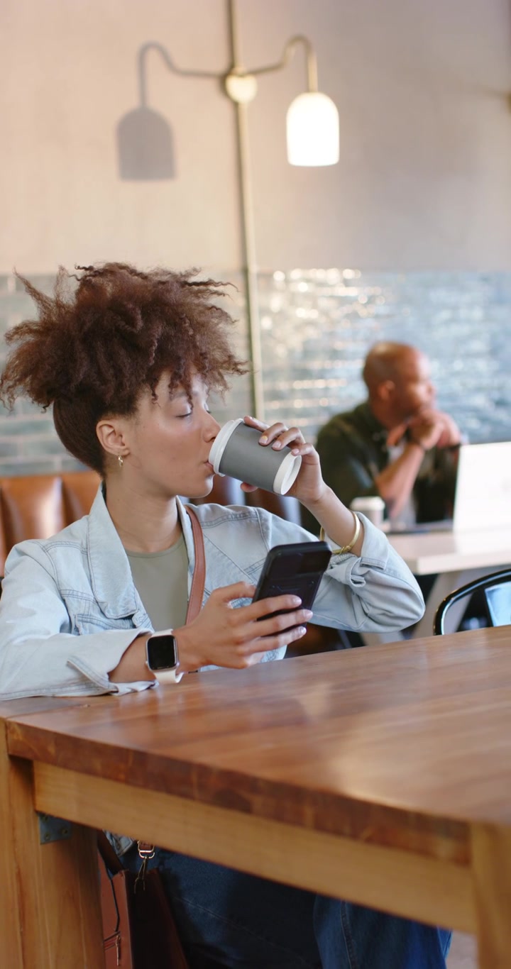 Video verticale: Donna afroamericana che legge messaggi sul telefono, sorseggiando da una tazza da asporto a un tavolo di legno