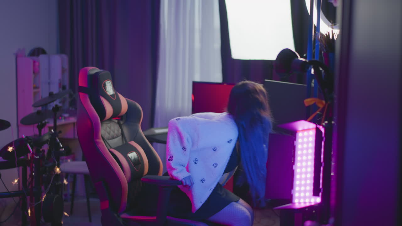 Female artist in fluffy sweater enters neon lit studio adjusts chair at desk with computer monitors ring light and white phone then sits under ambient pink purple glow before starting work