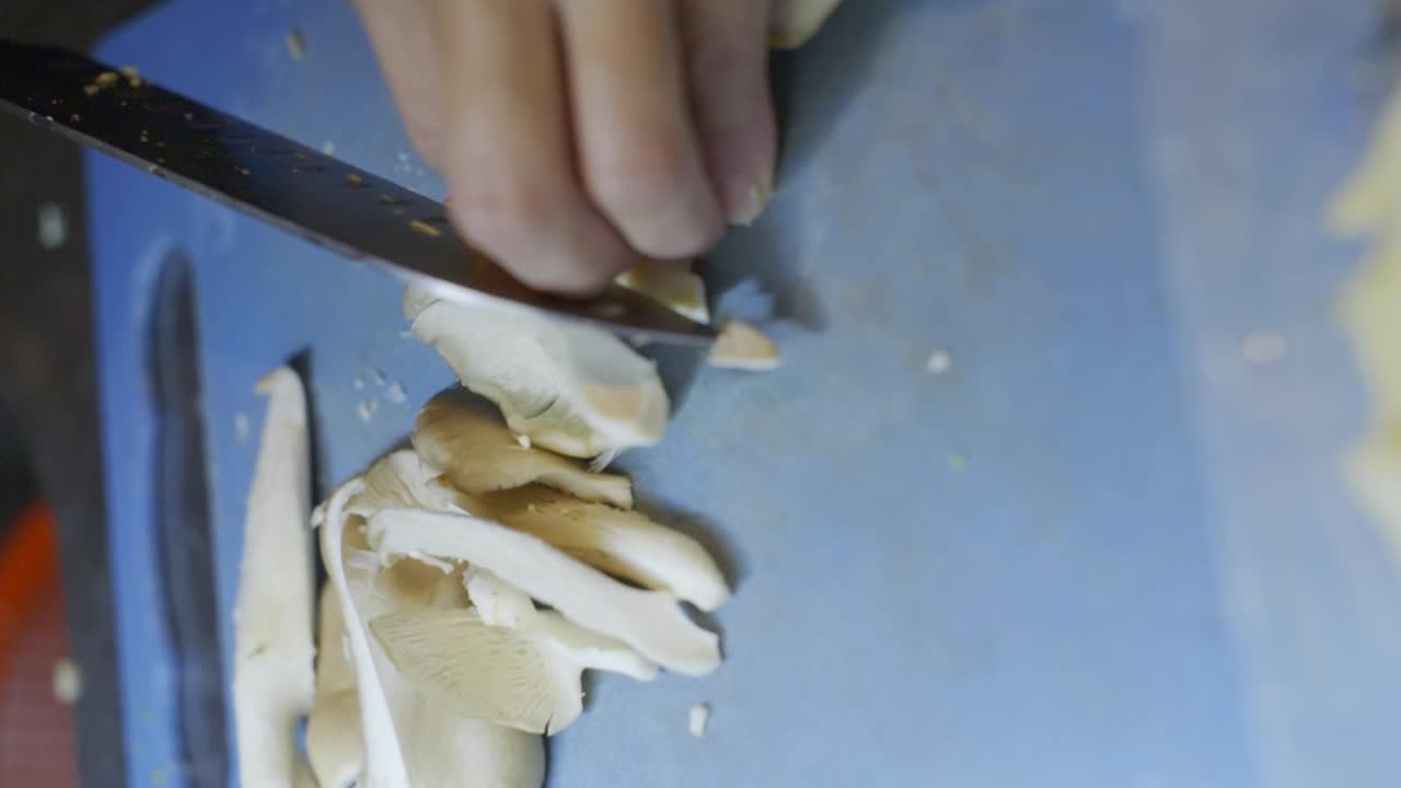 Knife used to cut mushroom into long slices on blue cutting board, filmed as vertical closeup handheld slow motion style