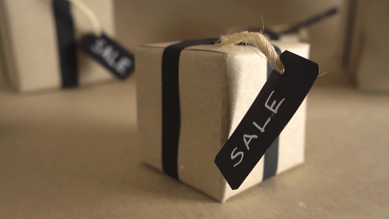 Enkel gaveeske innpakket i brunt papir med et svart bånd og en salgsetikett, som roterer i et reklamekonsept for julehandel, med grunn dybdeskarphet og myk bakgrunn.