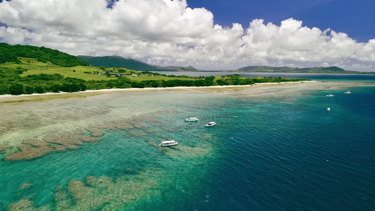 Stunning, static aerial shot of a pristine tropical paradise. Lush green hills meet a calm turquoise bay where boats are anchored over a visible coral reef under a blue sky with fluffy clouds