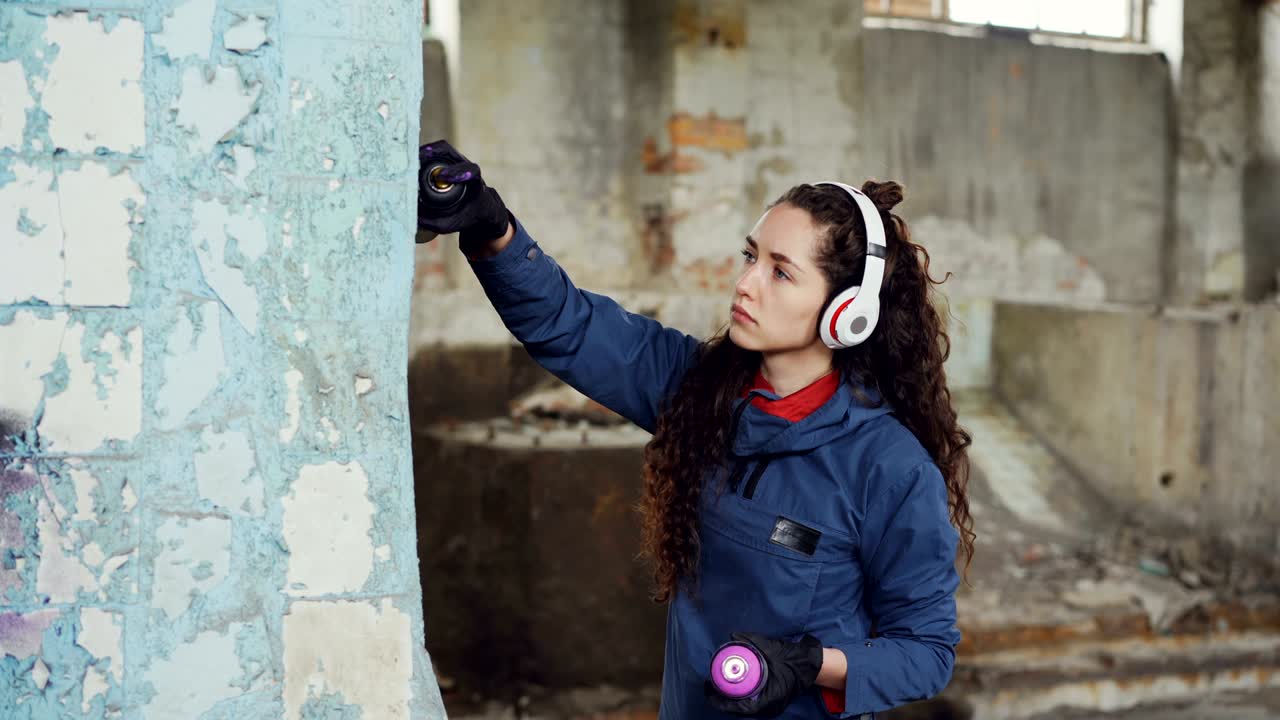 Creative young woman graffiti painter is using paint spray to decorate ruined pillar inside old empty warehouse. Girl is listening to music through wireless headphones.