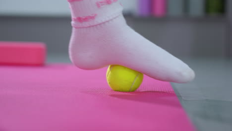 close-up of person leg in socks rolling a yellow tennis ball under foot on a pink mat, focused on muscle relaxation and foot massage techniques