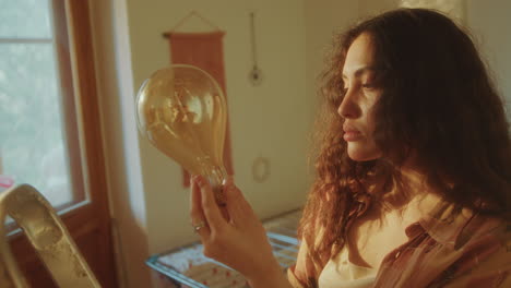 young woman standing on stepladder, examining light bulb in warmly-lit room