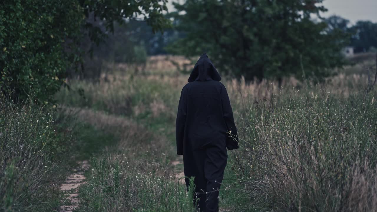 Backside view of witch on the field. Black death goes slowly away along the path on nature background.