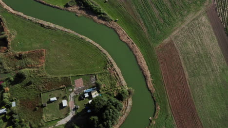 Aerial-top-down-shot-of-a-calm-curvy-river-among-the-fields-and-farms-of-a-small-village