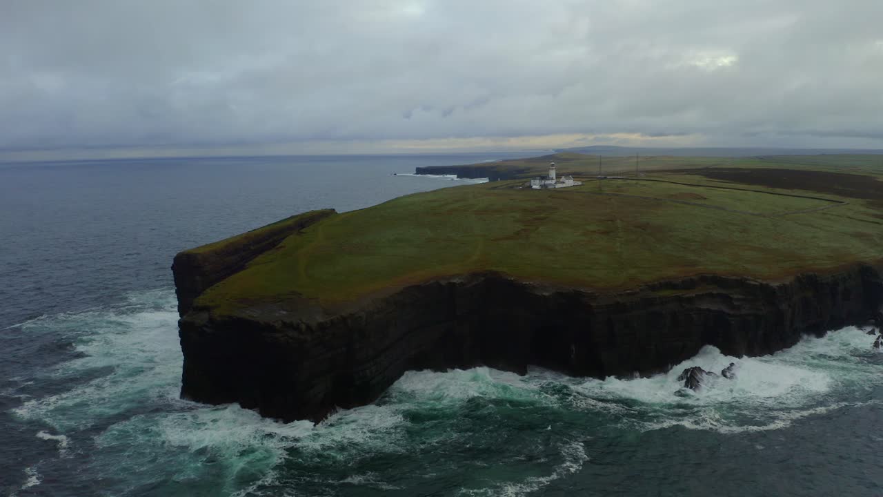 pan aérea muestra acantilados de cabeza de bucle y faro desde un ángulo de mar, con dramáticos cielos nublados y poderosas olas