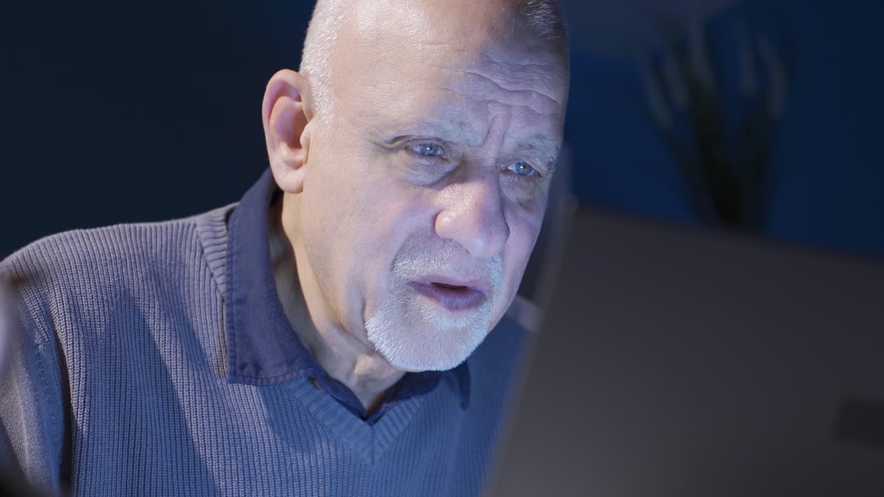 Old man looking at laptop at night working seriously and focused.
