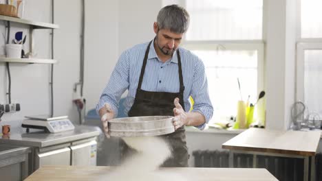 guapo panadero tamizando la harina a través de un tamiz en la cocina. cerca, en interiores, en cámara lenta