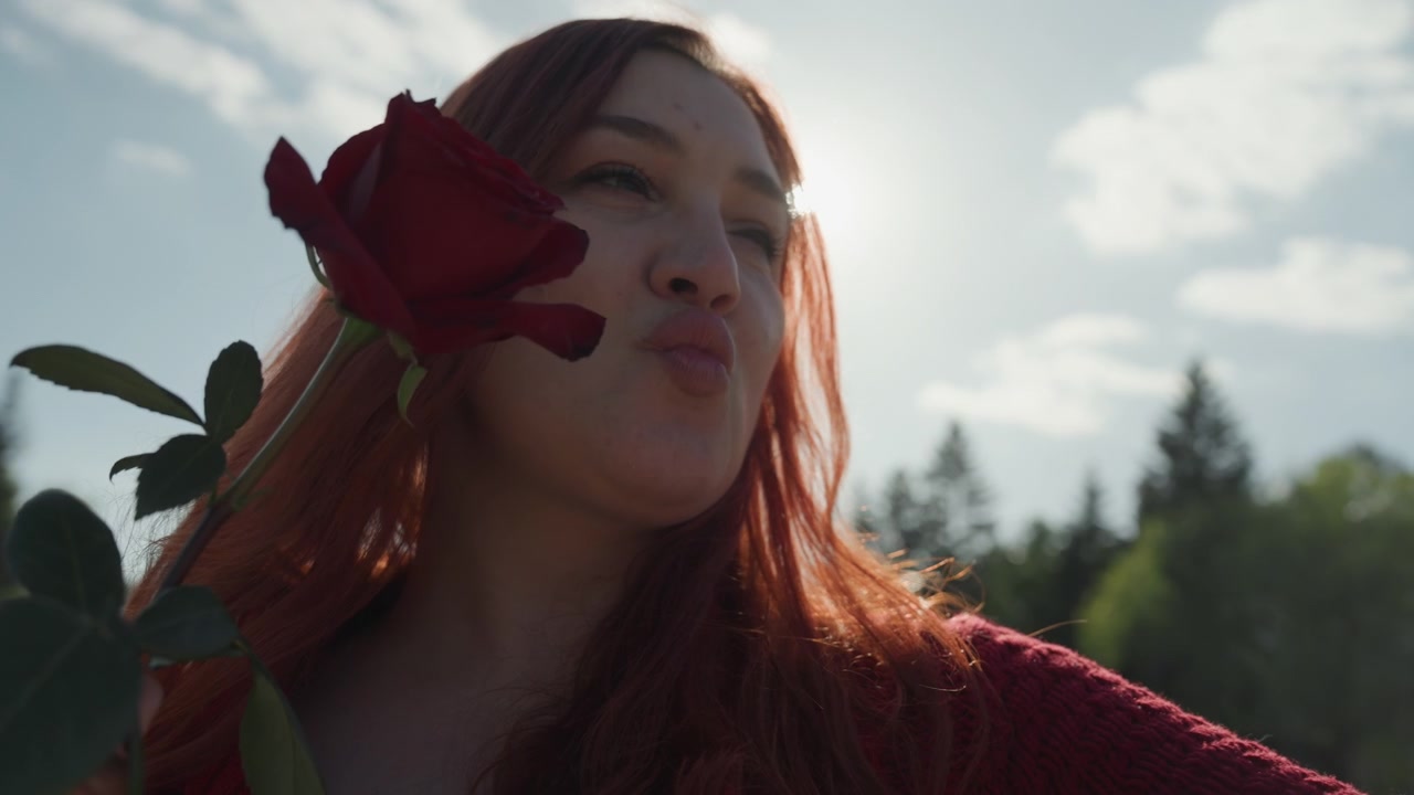 Primo piano di una donna che tiene e annusa una rosa rossa all'aperto, sotto la luce solare intensa con alberi verdi sullo sfondo. La donna indossa un maglione lavorato a maglia color bordeaux e interagisce delicatamente con il fiore. La luce del sole crea...