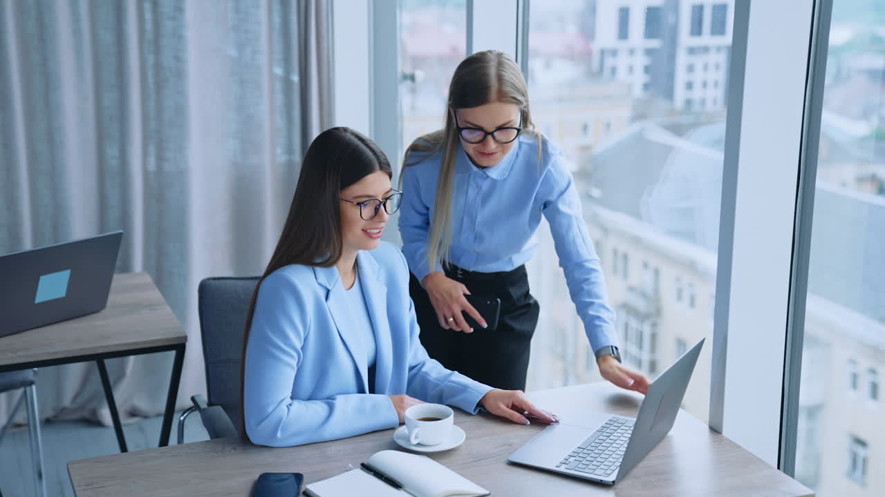 Two female colleagues collaborating to gain the best results at work. Young employees looking at laptop screen talking and smiling. Cityscape at backdrop in blur.