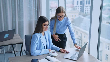 two female colleagues collaborating to gain the best results at work. young employees looking at laptop screen talking and smiling. cityscape at backdrop in blur.