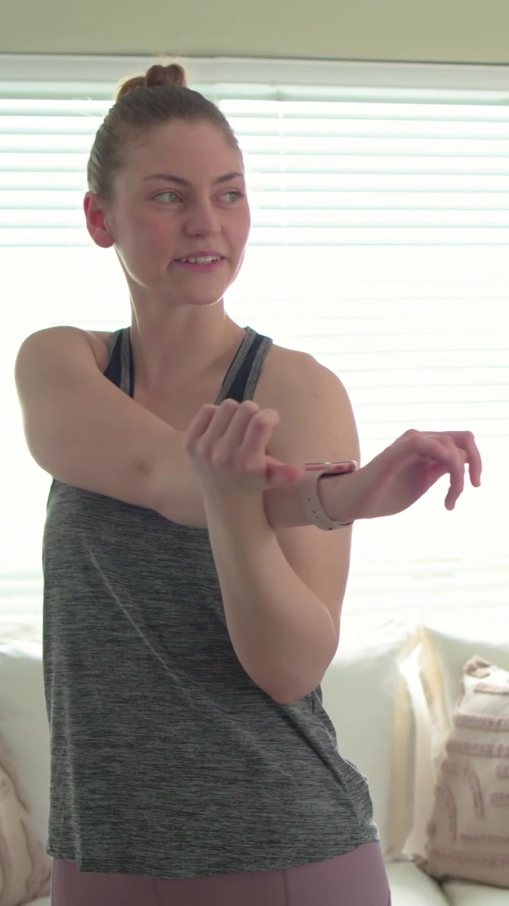 Video verticale: Donna che fa stretching in tenuta sportiva, controllando il tracker prima del riscaldamento a casa, con le tende.