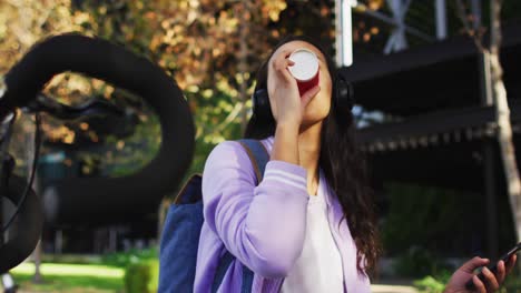 asian woman wearing headphones drinking coffee while sitting in the park