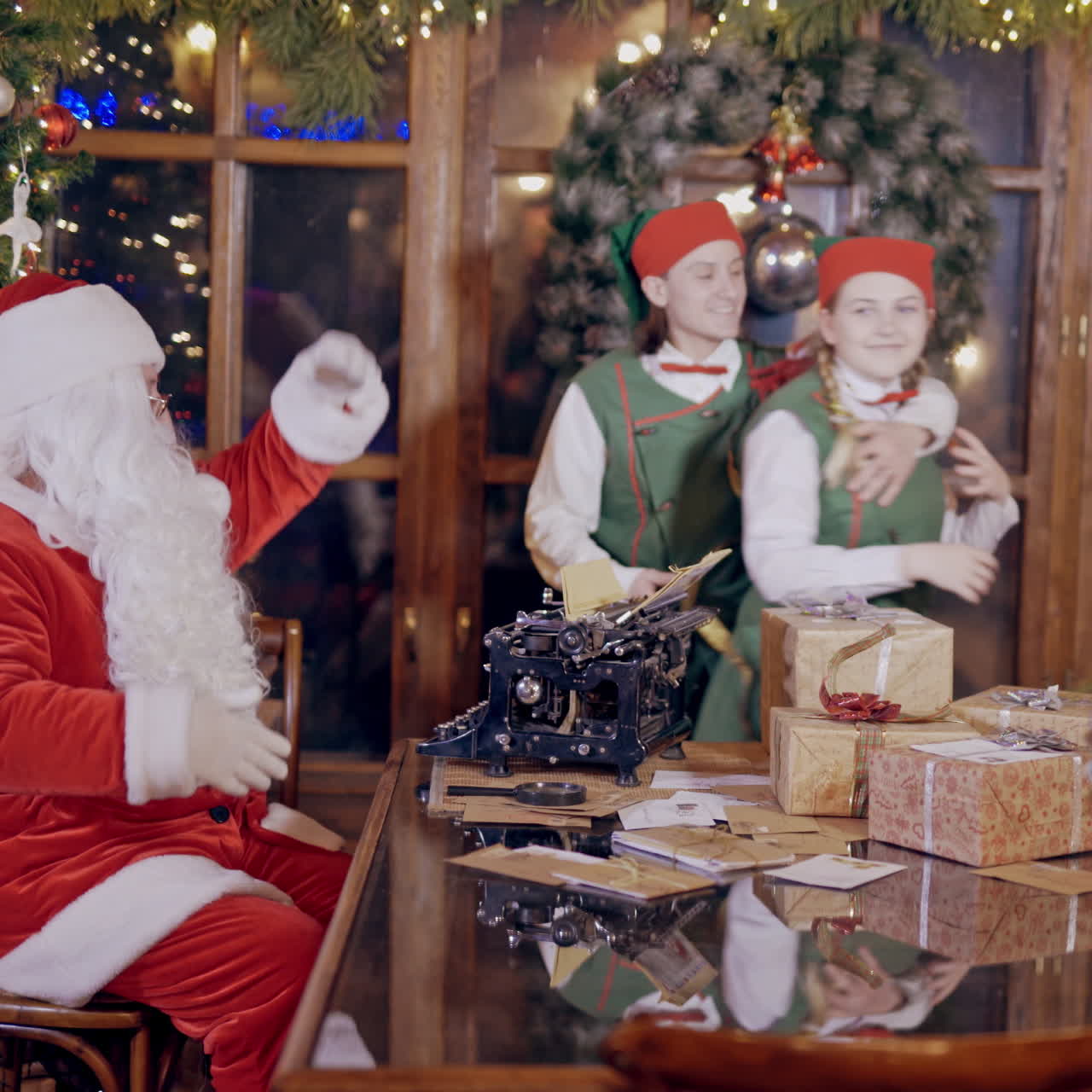Two merry elves in costumes are dancing together and Santa shows them movements by his hands. Santa Claus is sitting at the table with many gifts and letters on the background of elves.