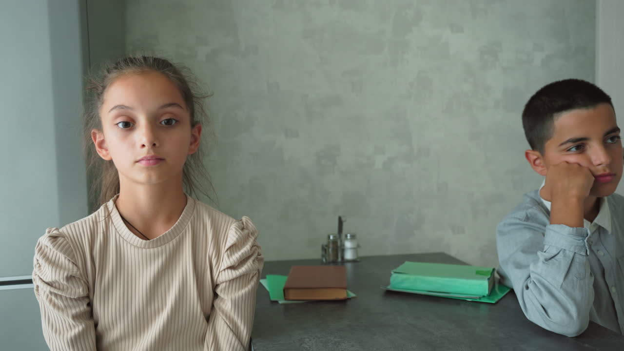 Girl with eyes and boy resting face on hand sit apart in silent modern room, both showing signs of boredom, stress, and disconnect as green folders and brown book lie on table between them