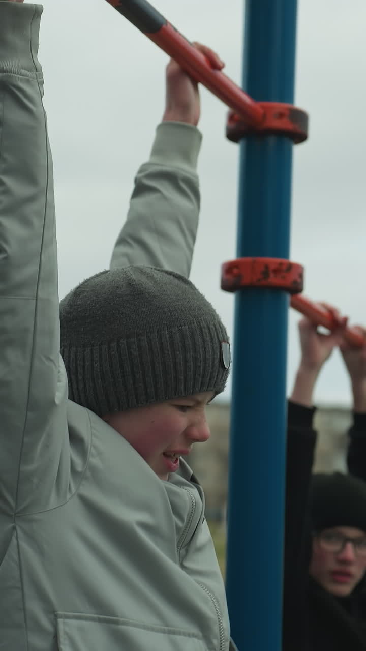 두 소년은 야외 체육관에서 풀업 바에서 운동을 하고 있으며, 회색 스웨터를 입은 소년은 계속하기 위해 고군분투하고 있으며, 검은 스웨터을 입은 소년이 멈추고 그를 쳐다보고 있습니다.