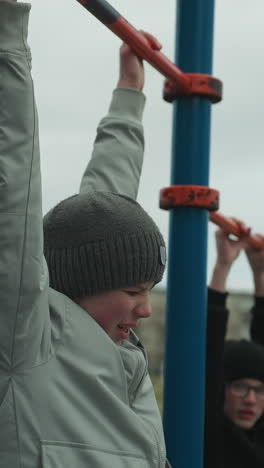 dos chicos están trabajando en barras de tracción en un área de gimnasio al aire libre, el chico con el suéter gris lucha por continuar, mientras que el chico en el suéter negro se detiene y lo mira