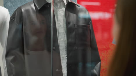 mannequin dressed in black leather jacket over checkered shirt displayed in retail store window, with subtle reflection of woman in glass, bold red and yellow background