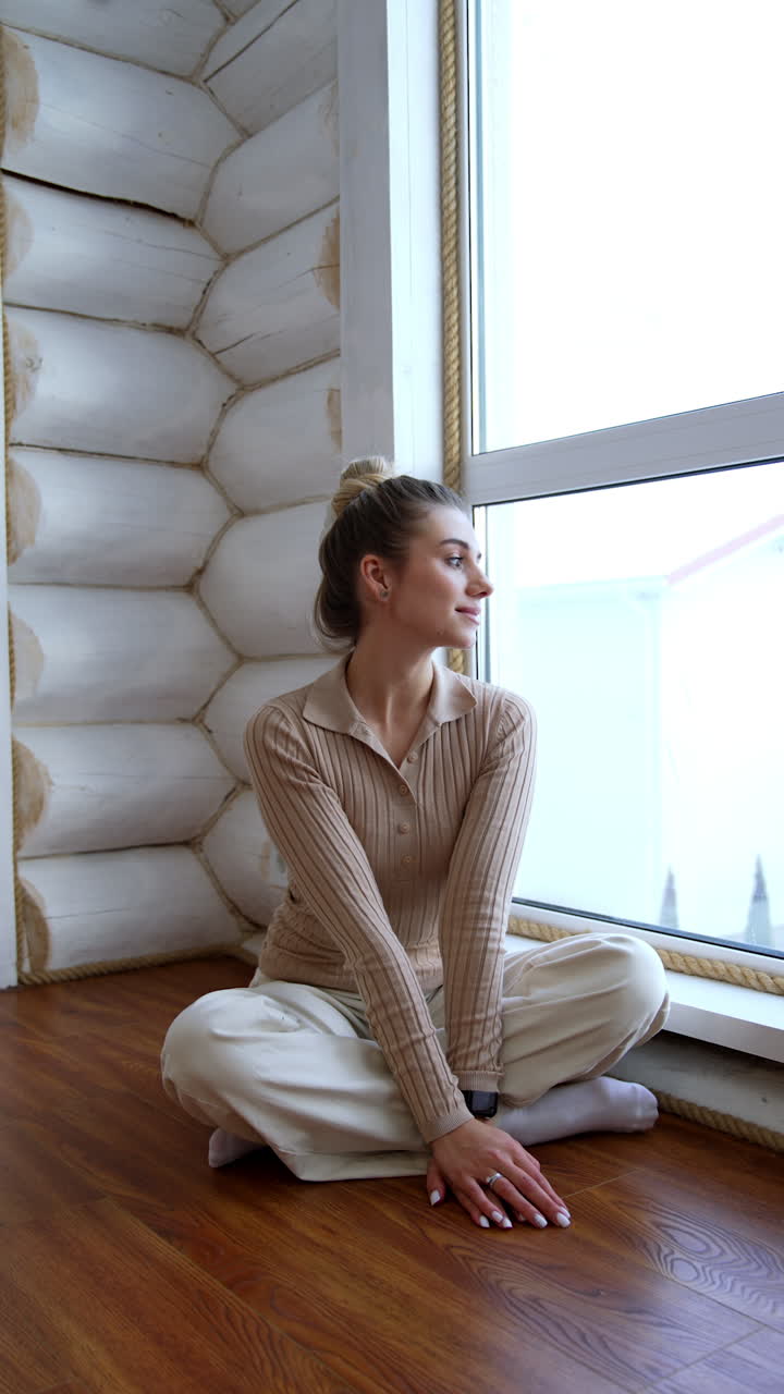 Beautiful Caucasian woman sits on the floor at the window. Relaxed positive lady looks outside. Vertical video.