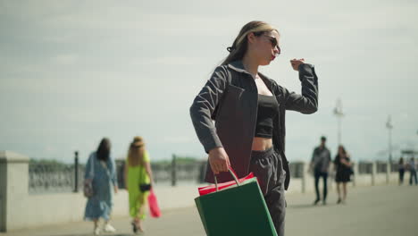 dama girando mientras baila y haciendo movimientos con su mano, sosteniendo bolsas de compras rojas, menta y verdes en su mano derecha, vista borrosa de personas caminando en el fondo