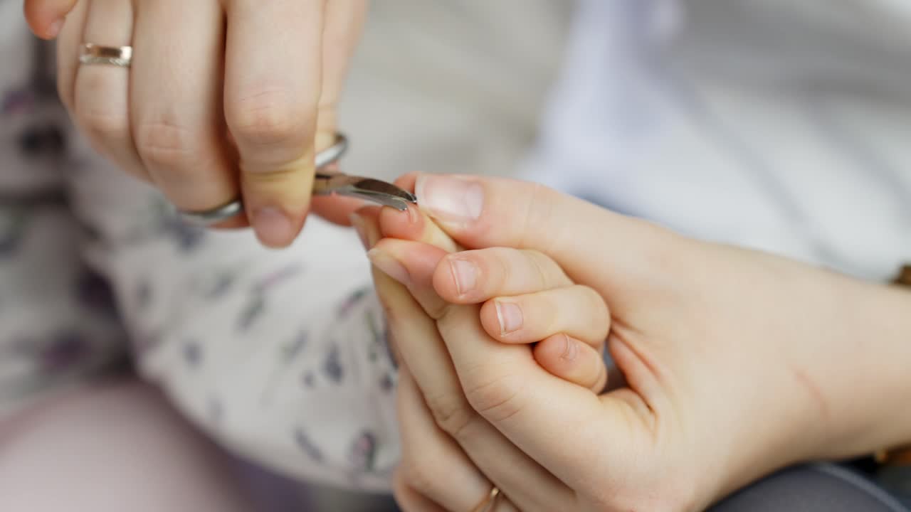 An adult delicately trims a young child’s fingernails using small scissors. This close-up moment captures care, hygiene, and gentle grooming in a calm and comfortable home environment.
