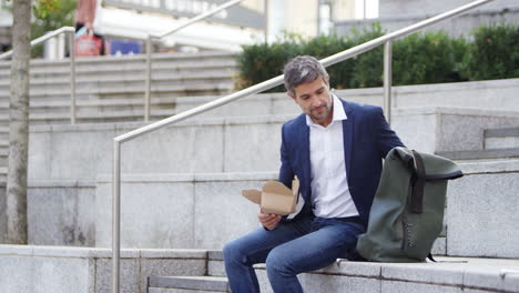 hombre de negocios sentado afuera en la pausa para el almuerzo comiendo comida para llevar de una caja de cartón reciclable sostenible