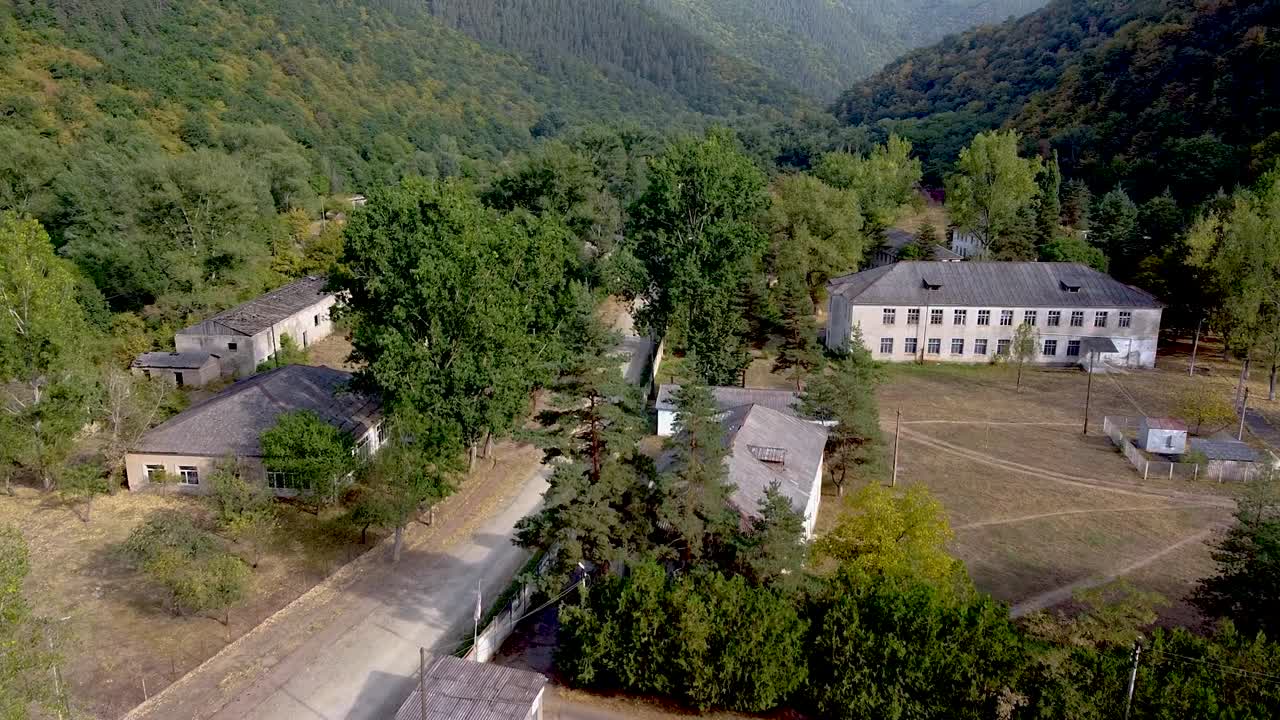 Experience an aerial view of an abandoned building complex nestled in a green valley, with tall trees and mountains in the background. Nature slowly reclaims the area