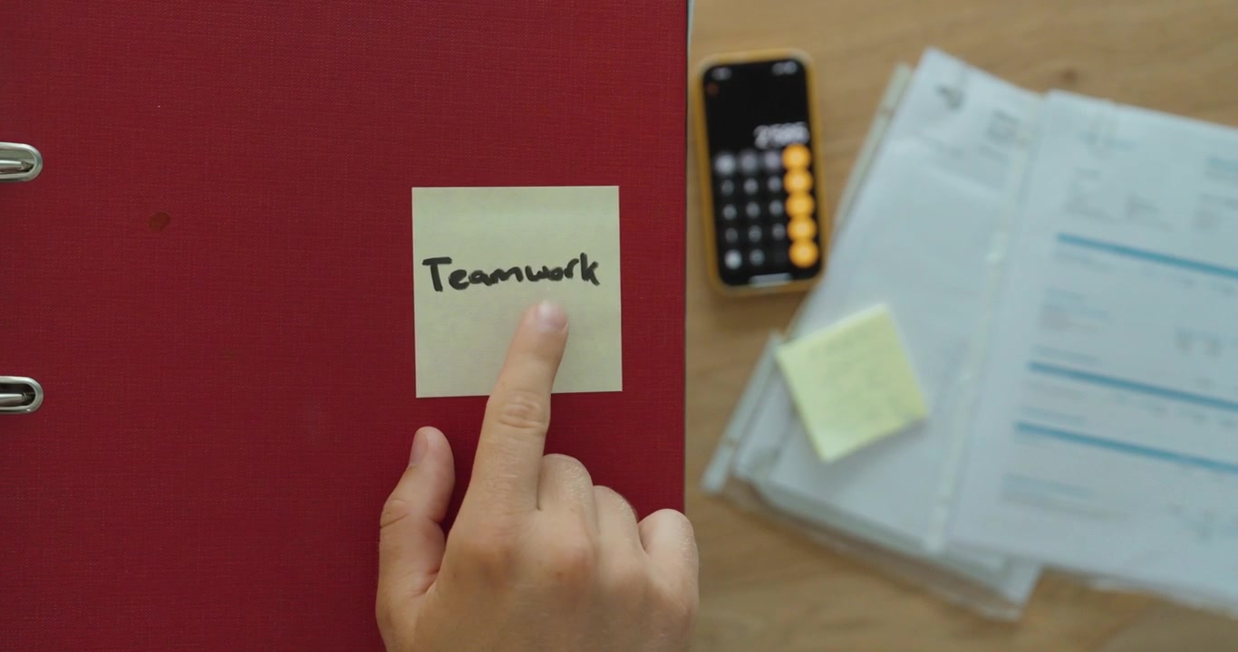 Pointing at a teamwork sticky note with business documents in the background. Ideal for collaboration, leadership, office productivity, and teamwork concepts.