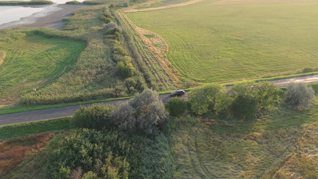 A single car travels along a deserted road through wide open green fields. The peaceful rural landscape is bathed in warm sunlight, creating a serene and tranquil scene