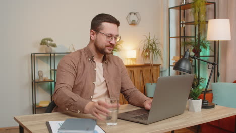 focused caucasian businessman sitting at desk drinking water while working on laptop in home office