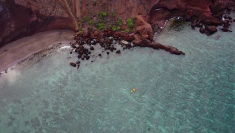 Stunning-aerial-view-of-tourists-rowing-in-a-colorful-kayak-in-tropical-Hawaiian-exotic-destination-with-turquoise-calm-ocean-surface-and-red-dirt-cliffs