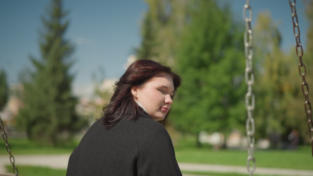 검은 옷을 입은 여성이 야외에서 빛 아래 머리카락을 조정하는 동안, 활기찬 초록색, 경로, 그리고 공공 공원에서 멀리 있는 나무와 함께