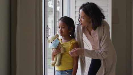 Hispanic-mother-and-daughter-embracing-in-window-holding-teddy-bear-with-face-mask