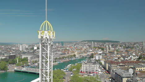 Un-Dron-Aéreo-Sobrevoló-La-Torre-De-Caída-Libre-Del-Parque-De-Atracciones-Con-La-Ciudad-Y-El-Lago-De-Zúrich,-Suiza-En-El-Fondo-Durante-El-Zürichfest