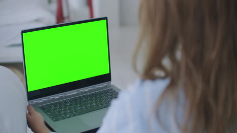 Junge-Frau-Arbeitet-Zu-Hause-An-Einem-Laptop-Computer-Mit-Grünem-Mock-up-Bildschirm.-Sie-Sitzt-Auf-Einer-Couch-In-Seinem-Gemütlichen-Wohnzimmer.-Kameraaufnahme-über-Die-Schulter