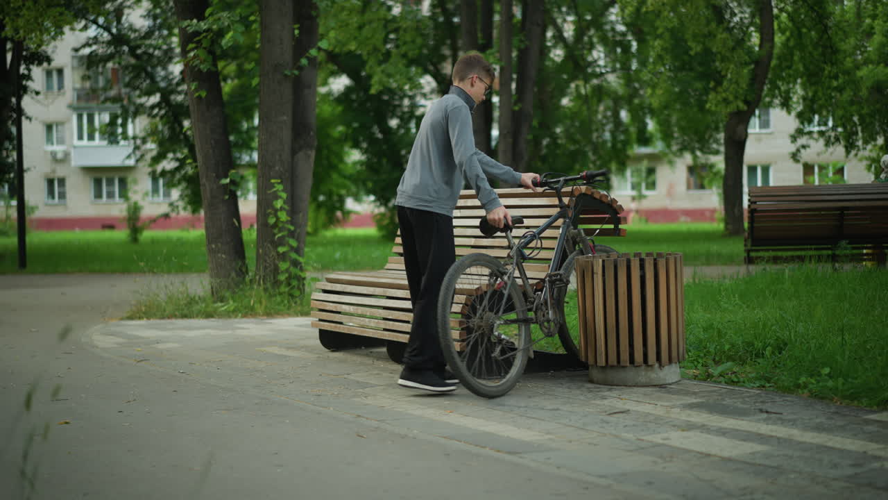 소년은 평화로운 공원을 통해 자전거를 타고 나무 벤치에 멈추고, 벤치와 근처의 나무 구조물 사이에 자전거을 주차하고, 다른 사람은 녹색으로 둘러싸인 먼 벤치에서 앉아 있습니다.