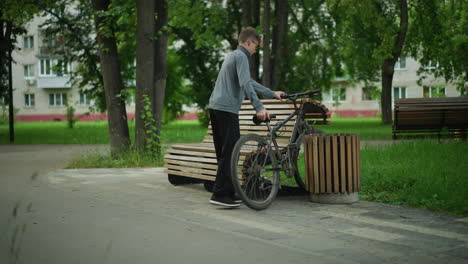 boy rides bicycle through peaceful park, stops by wooden bench, parks bike between bench and nearby wooden structure, another person sitting on distant bench surrounded by greenery