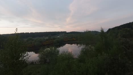 Still-lake-in-a-country-park-at-dusk-on-a-sunset-evening-establishing-revealing-aerial-shot