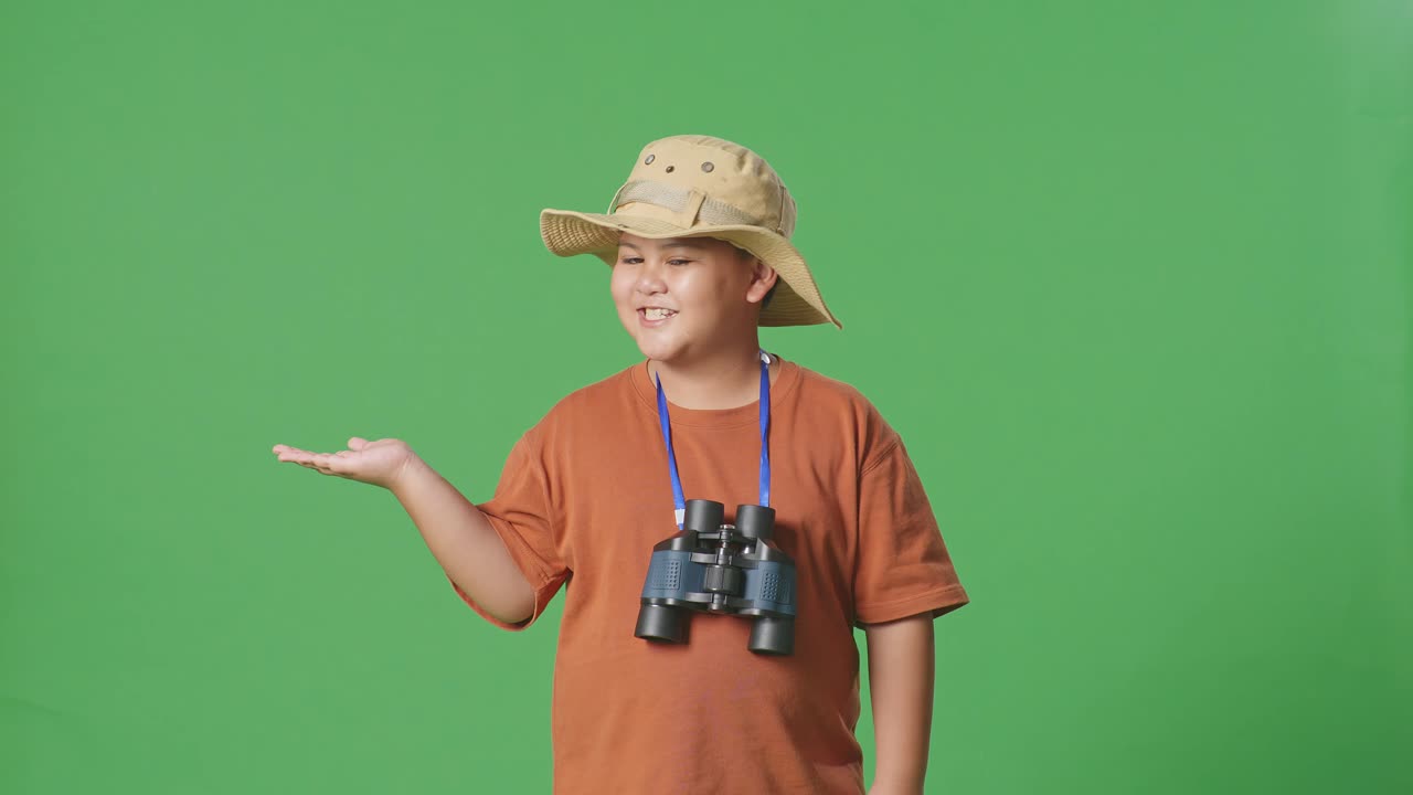 모자와 안경을 쓴 아시아 관광 소년이 미소 짓고 을 가리키며 녹색 스크린 배경에 서 있습니다. 소년 연구자는 무언가를 조사합니다.