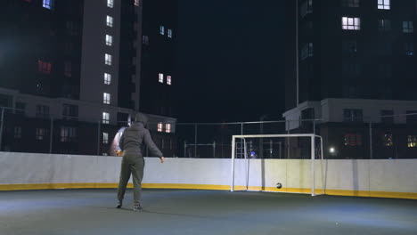 man kicks soccer ball toward goalpost under artificial night lighting on outdoor court, with urban background and partial view of person walking behind fence