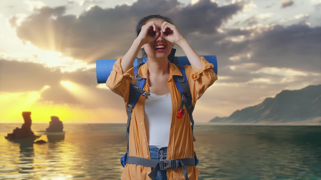 Senderista asiática sonriendo con una mochila de montañismo, haciendo el gesto de los prismáticos y mirando alrededor mientras está de pie en la playa durante la puesta de sol
