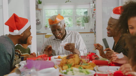 Familia-Multigeneracional-Con-Sombreros-De-Papel-Leyendo-Chistes-De-Galletas-Navideñas-Mientras-Comen-Juntos