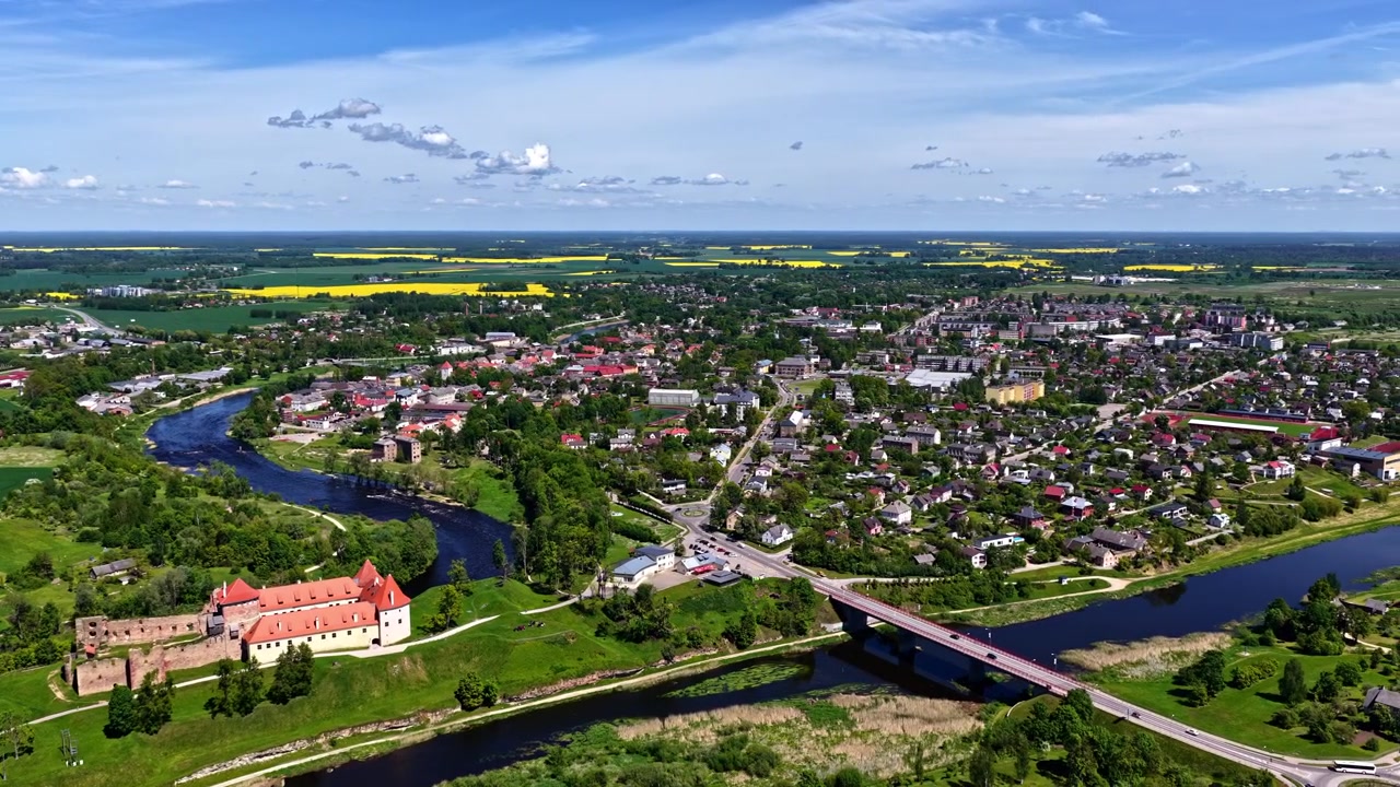 Toma panorámica con dron del histórico Castillo de Bauska, el río y la ciudad bajo un cielo azul brillante.