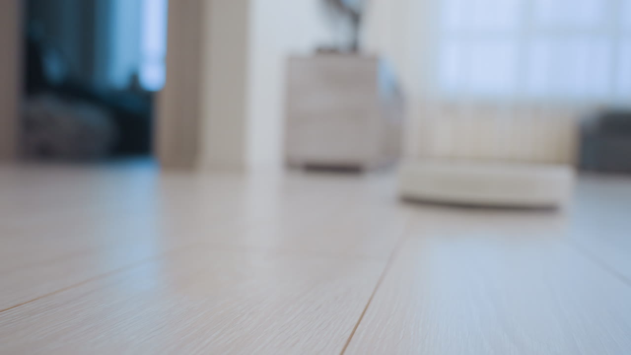 Close up of robot vacuum cleaner moving across wooden floor turning toward bright light with blurred background, showing smart cleaning technology